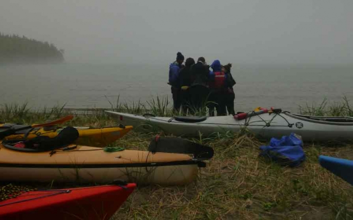 a group of outward bound students huddle in a circle on the shoreline of a lake. kayaks rest on the shore. 
